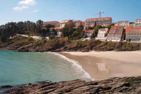 La **playa Caneliñas** se asoma entre rocas y vegetación, con edificios residenciales en la colina y el mar turquesa.