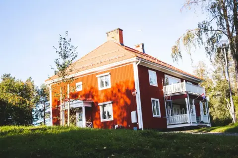 Rött hus med vita knutar och veranda, omgivet av grönska. Ett charmigt vandrarhem i Jokkmokk som bjuder in till en naturnära vistelse.