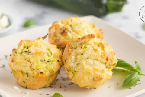 Drei goldbraune Zucchini-Muffins mit Kräutern und Käse auf einem Teller, garniert mit Basilikum.