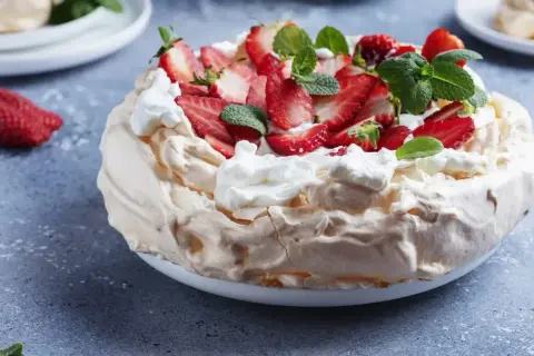 Puszysta beza Pavlova ozdobiona bitą śmietaną, świeżymi truskawkami i listkami mięty. Idealna na letnie popołudnie.