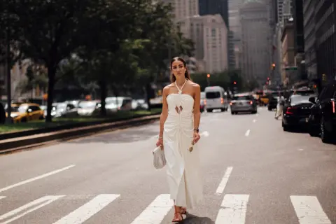 Eine Frau in einem weißen Kleid überquert eine Straße in New York. Sie trägt rote Sandalen und hält eine Sonnenbrille und eine Handtasche. Kleine Frauen sind hier nicht zu sehen.