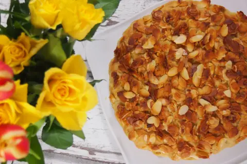 Bienenstich mandelkruste ohne backen, verziert mit gerösteten Mandelsplittern. Daneben ein Strauß gelber Rosen und Tulpen.