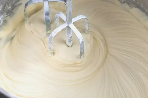Mieszadła miksera obracają się w gładkim, kremowym **kremie z mascarpone i białej czekolady**, tworząc wiry.