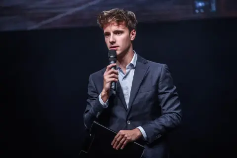 Młody mężczyzna w garniturze przemawia do mikrofonu, zastanawiając się, ile zarabia przeciętny aktor.