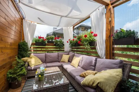 Przytulne mieszkanie z ogródkiem, z wygodną sofą i stolikiem kawowym pod zadaszeniem, otoczone kwitnącymi pelargoniami.