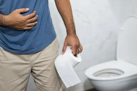 Mężczyzna trzymający papier toaletowy, z ręką na brzuchu, w łazience. Prawdopodobnie doświadcza biegunki po alkoholu.