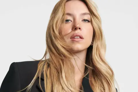 Sydney Sweeney, con su cabello rubio ondulado y un blazer negro, mira directamente a la cámara con una expresión cautivadora.