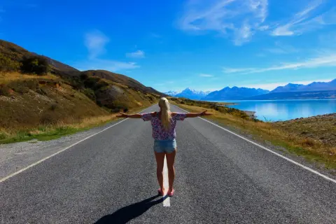 Frau mit ausgebreiteten Armen auf einer Straße, die zu schneebedeckten Bergen und einem blauen See führt. Ein Traum von Work and Travel Weltreise.