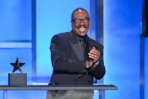 Eddie Murphy, känd för sina roliga filmer, ler och klappar händerna på scenen bredvid en stjärnstatyett.
