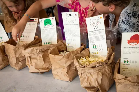 Barn plockar popcorn ur papperspåsar. En påse med popcorn med kolasås syns tydligt.