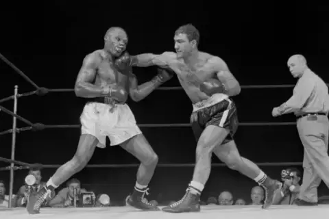 Two boxers in a ring, one landing a punch. The boxer on the right, with a powerful Rocky Marciano physique, is throwing a jab.