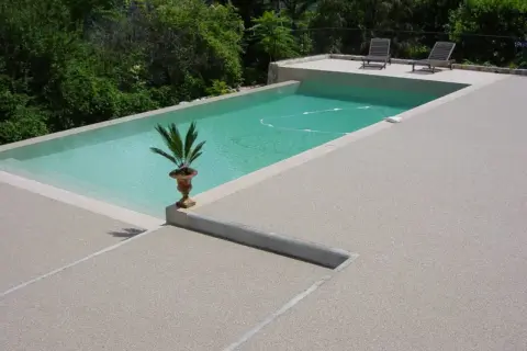 Piscina con bordo in cemento e pavimentazioni in cemento a vista, circondata da vegetazione lussureggiante.