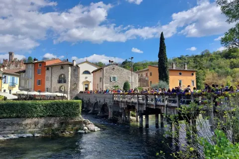 Un pittoresco borghetto mantovano con case colorate, un ponte di legno affollato di gente e un fiume che scorre sotto un cielo azzurro con nuvole bianche.