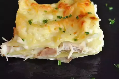 Une part de lasagnes poulet champignons, gratinée et garnie de persil frais, sur une assiette noire.