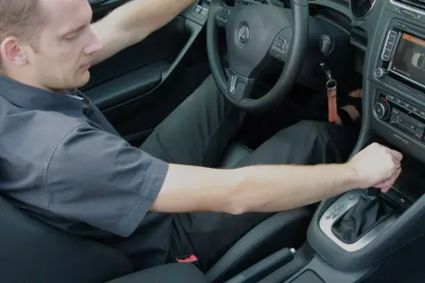 Mężczyzna w czarnej koszuli i spodniach, siedząc za kierownicą samochodu z automatyczną skrzynią biegów, czeka na rozwinięcie się korka.