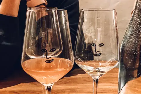 Dwa kieliszki z zabawnymi buźkami na drewnianym stole w wine barze w Gdańsku. Jeden z nich zawiera różowe wino.