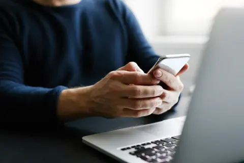 Mężczyzna trzyma telefon, szukając informacji, jak przenieść kontakty na kartę SIM. Obok leży laptop.