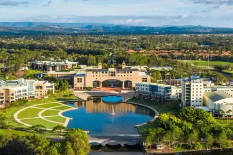 Weite Luftaufnahme des Campus der Bond University mit einem zentralen See, gepflegten Grünflächen und modernen Gebäuden, umgeben von Bäumen und Hügeln.