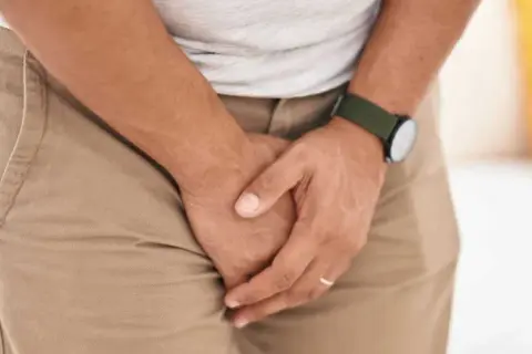 Mężczyzna w beżowych spodniach i szarej koszulce obejmuje się rękami w kroku, sugerując problem z nietrzymaniem moczu u mężczyzn.