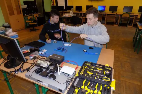 Dwóch uczniów ćwiczy technikę informatyczną, montując kable sieciowe. Na stole sprzęt i narzędzia.