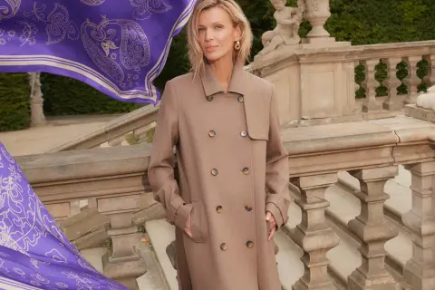 Elegancka kobieta w beżowym płaszczu, obok niej powiewa fioletowy szal z wzorem. Zastanawiasz się, jaki kolor pasuje do fioletu? Beż jest doskonałym wyborem!