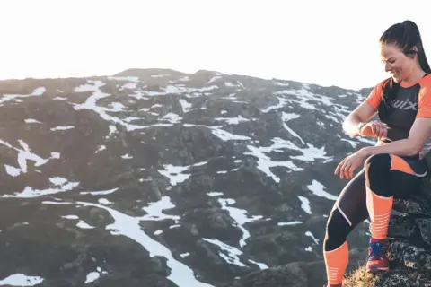 Une femme en tenue de sport s'assoit sur des rochers, regardant sa montre, prête à trouver un trail dans un paysage montagneux enneigé.