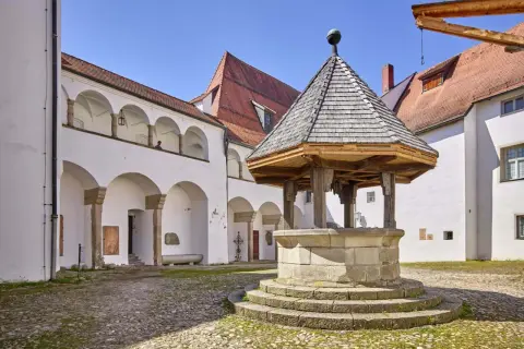 Historischer Brunnen im Innenhof des Oberhaus Passau. Weiße Arkadenbögen und rote Ziegeldächer unter blauem Himmel.