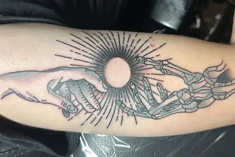 Ein Tattoo zeigt eine menschliche Hand und eine Skeletthand, die sich auf einen hellen Kreis zubewegen. Die Haut ist leicht gerötet, als ob sie gerade erst aufgerissen wurde, um das Kunstwerk zu enthüllen.