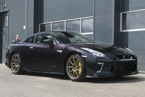 Ciemny Nissan R35 z brązowymi felgami i żółtymi zaciskami hamulcowymi stoi przed szarym budynkiem.