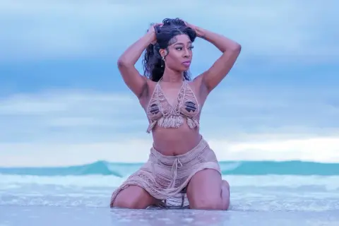 Modella in costume da bagno con frange, in ginocchio sulla riva del mare, con le mani tra i capelli.