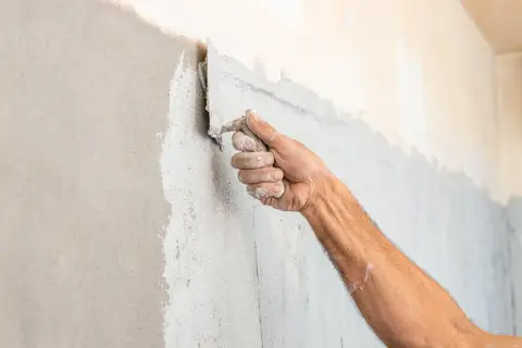 Mano che applica intonaco spugnato su una parete con una spatola. La texture è grezza e irregolare.