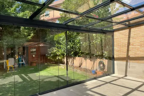 Modernos tejados para porches de cristal, con vistas a un jardín con caseta de madera y árboles frondosos.