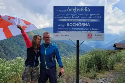 Para podróżników z Gruzji, pytanie "czy w Gruzji jest bezpiecznie" nabiera nowego znaczenia. Para z gruzińską flagą na tle górskiego krajobrazu i tablicy informującej o najwyżej położonej wsi w Europie.