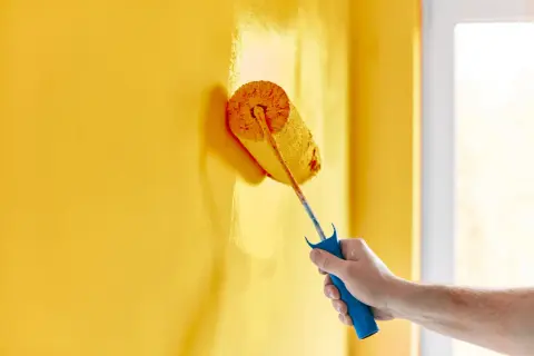 Mano che imbianca un muro giallo con un rullo. La temperatura ideale per imbiancare è tra i 10°C e i 25°C per una buona asciugatura.