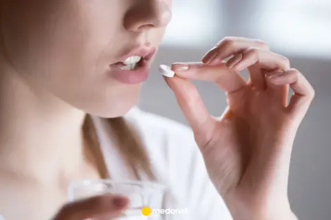 Kobieta bierze tabletkę clindamycin, trzymając ją między palcami. Obok widać szklankę z wodą.