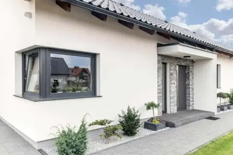 Nowoczesny dom z szarym, narożnym oknem ze słupkiem, kamienną elewacją przy drzwiach i zadbanym ogrodem.