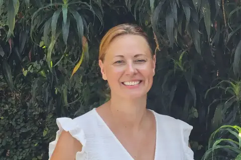 Vrolijke vrouw met blonde haren, lachend in een witte jurk, tegen een achtergrond van weelderig groen. Arjan van Dam zou trots zijn op deze foto.