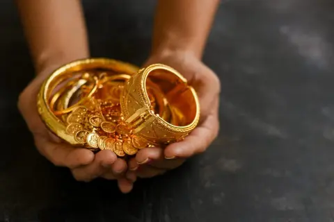 Manos sostienen un tesoro de joyas de oro, pulseras y monedas, un reflejo de las curiosidades del oro y su valor eterno.