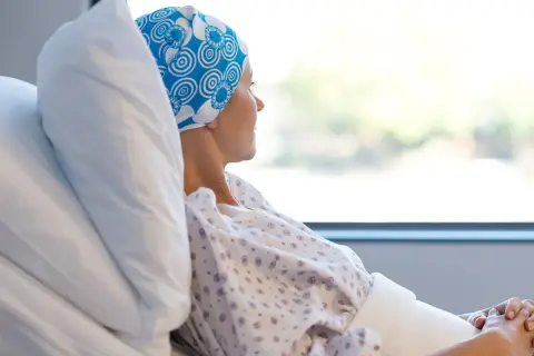 Kobieta w chustce na głowie leży w szpitalnym łóżku, patrząc przez okno. Czasem choroba nowotworowa może powodować dziwny zapach od osoby chorej na raka.