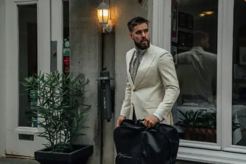 Mann im cremefarbenen Anzug mit gemustertem Hemd und Krawatte, trägt eine schwarze Kleidersacktasche. Ein lässiges Outfit für unterwegs.