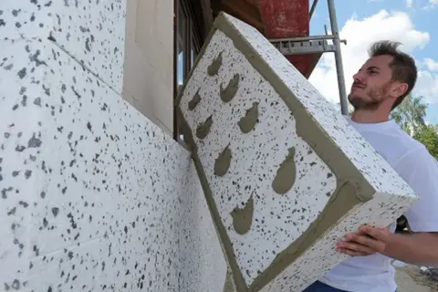 Budowlaniec montuje płytę z polistyrenu na ścianie. Materiał izolacyjny ma szary granulat.