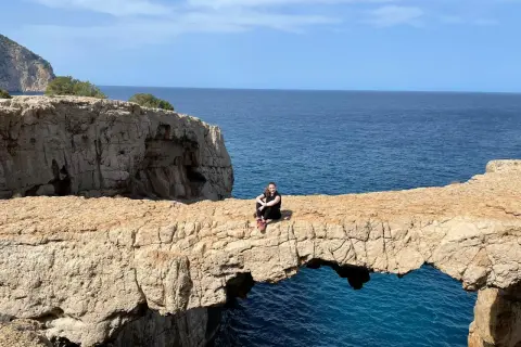 Una mujer sentada en un arco de roca natural, el "cala columpio ibiza", con el mar azul de fondo.
