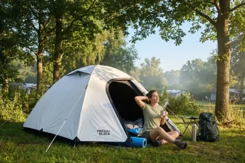 Une femme savoure un café matinal près de sa tente, profitant du calme pour bien dormir en tente. Un sac à dos et un thermos sont à proximité.