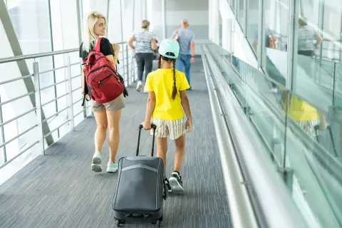 Młoda kobieta z plecakiem i dziewczynka z walizką idą korytarzem lotniska. To może być ich pierwszy wyjazd nieletniego za granicę bez rodziców.