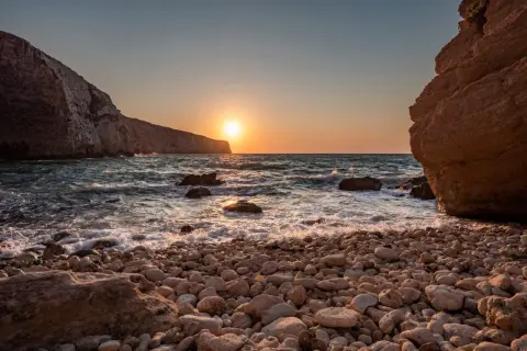 Malta zachód słońca nad kamienistą plażą. Słońce odbija się w falach, a klify tworzą malowniczą scenerię.