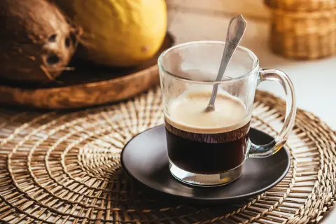 Tazza di caffè fumante su sottobicchiere intrecciato. Sullo sfondo, cocco e melone. Un caffè lungo ha più caffeina per iniziare la giornata.