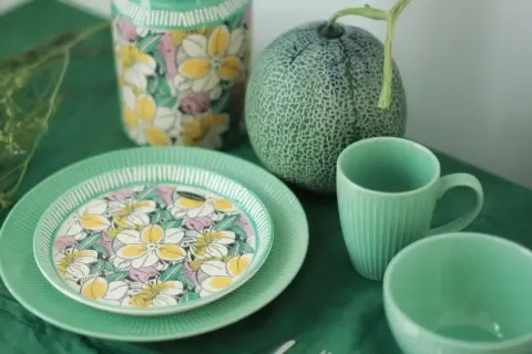 Assiette en bone china porcelaine ornée de fleurs colorées, à côté d'une tasse et d'un bol verts, avec un melon et un vase fleuri.