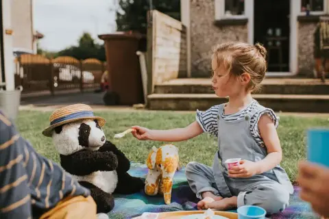 Dziewczynka karmi pluszową pandę w kapeluszu. Zabawa w piknik to świetna okazja do rozwiązywania krótkich zagadek.