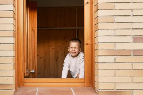 Niño sonriente asoma por una ventana de madera recién instalada.