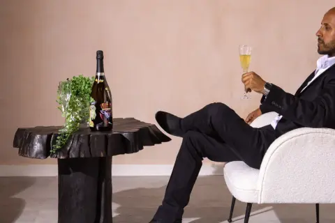 Mann in schwarzem Anzug genießt ein Glas Sekt neben einer Flasche, die wie der teuerster Sekt der Welt aussieht.